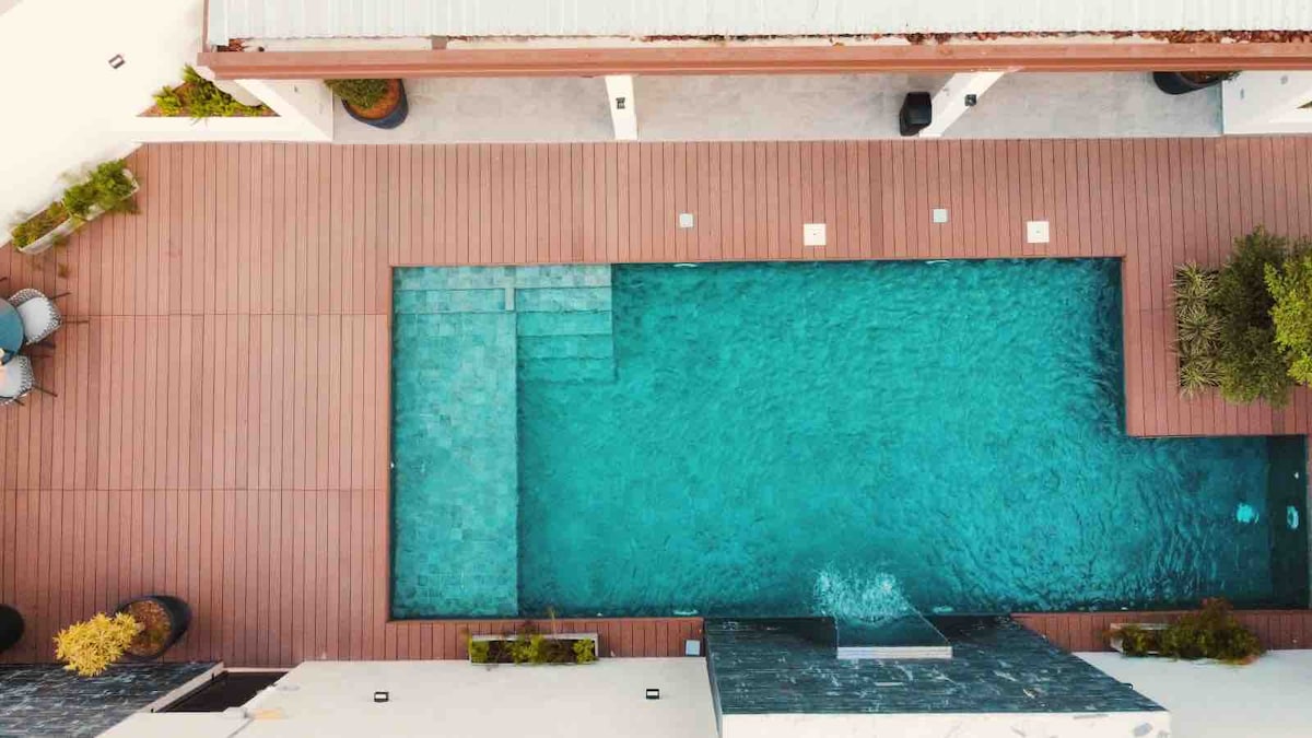 An aerial view captures a tranquil pool surrounded by wooden decking. The pool features a distinct shape with varying depths, and adjacent areas are accented by lush greenery and modern landscaping. Sunlight reflects off the clear water, creating a serene atmosphere.