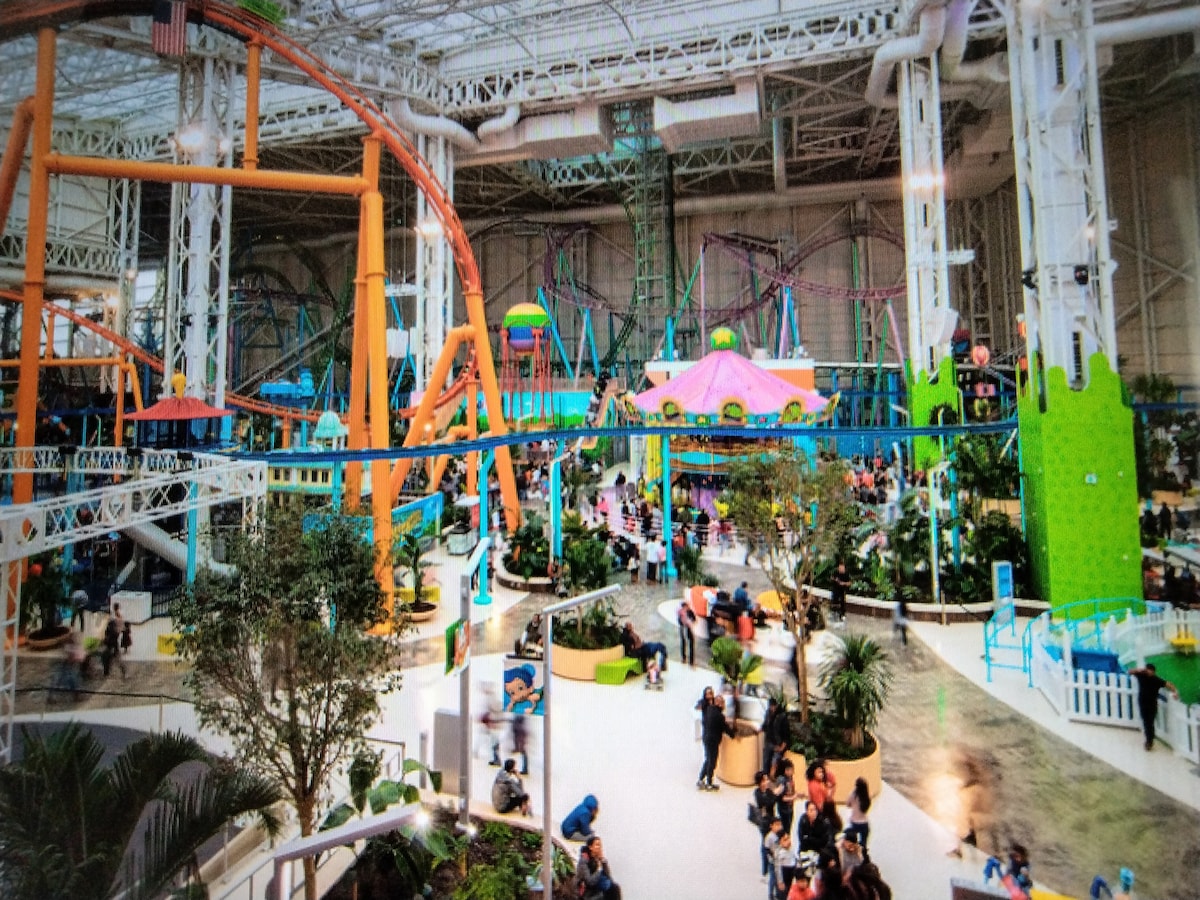 A colorful indoor amusement park is filled with various rides and attractions, including roller coasters and vibrant structures. Lush greenery is interspersed throughout the spaces, while groups of visitors are seen enjoying the lively atmosphere.