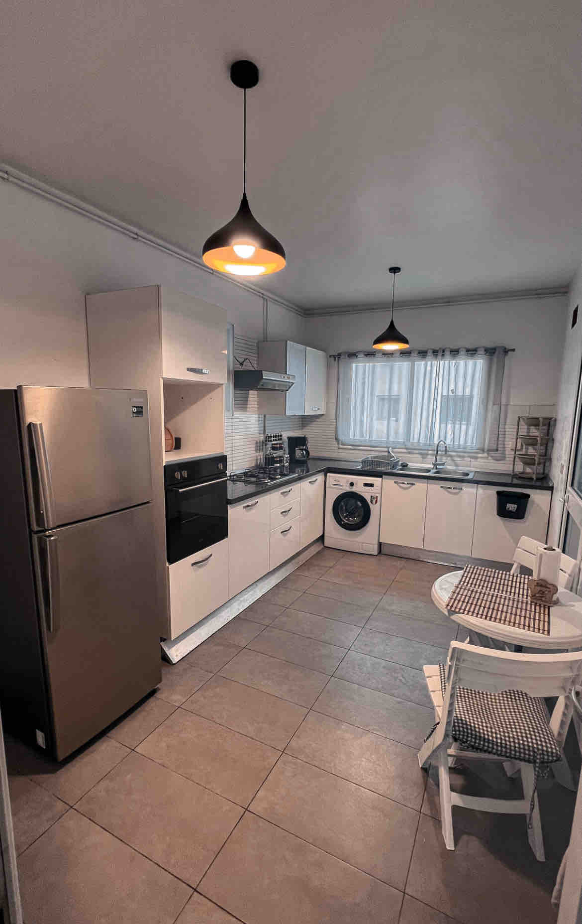 The kitchen features modern cabinetry and appliances, including a stainless steel refrigerator and oven. A washing machine is visible, alongside a small dining table with two chairs. Natural light filters through a window, brightening the spacious layout with neutral tones and tiled flooring.