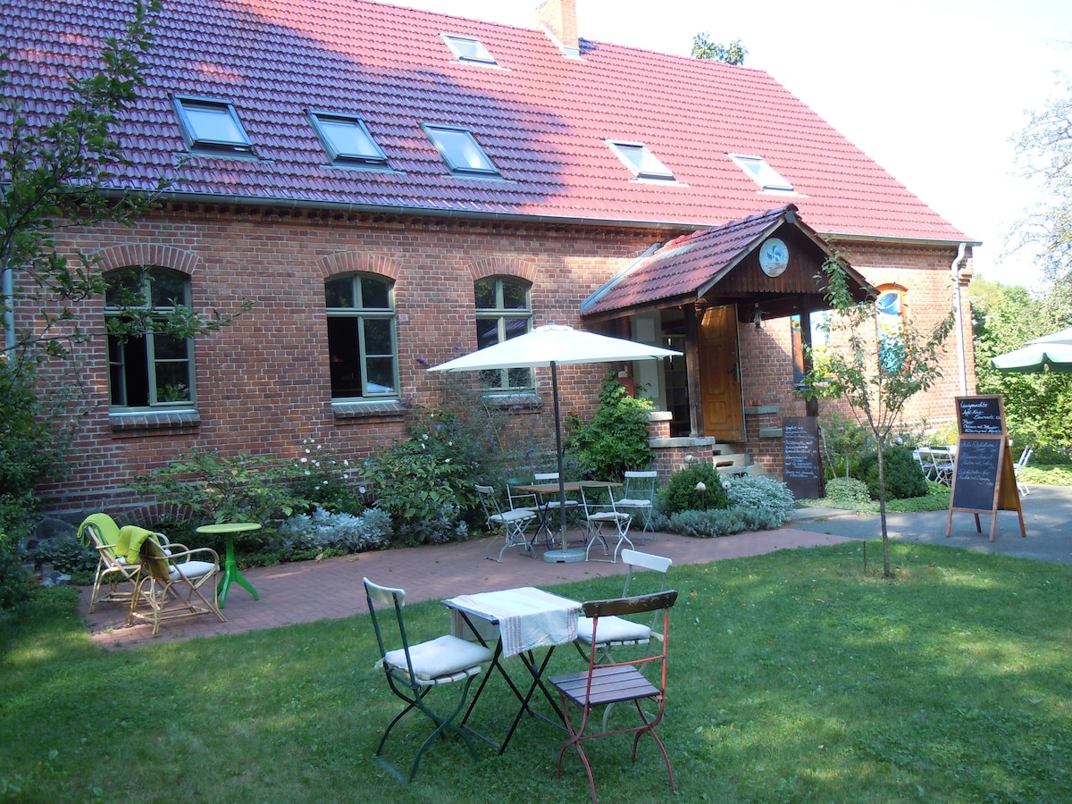 The charming brick exterior of a historic building is complemented by a red tiled roof. In the foreground, a small outdoor seating area features a table and chairs surrounded by lush greenery. An umbrella provides shade, enhancing the inviting garden atmosphere.