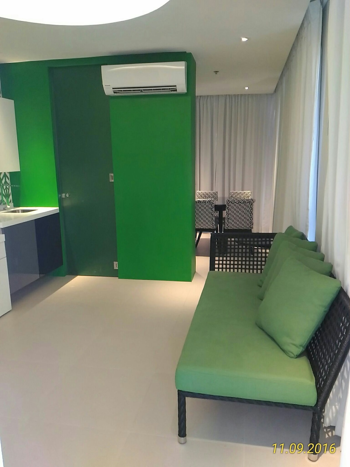 A bright living space is complemented by vibrant green walls, creating an energetic ambiance. A comfortable black sofa with green cushions offers seating, while a dining area with patterned chairs is positioned nearby. The room is illuminated by natural light through large curtains.