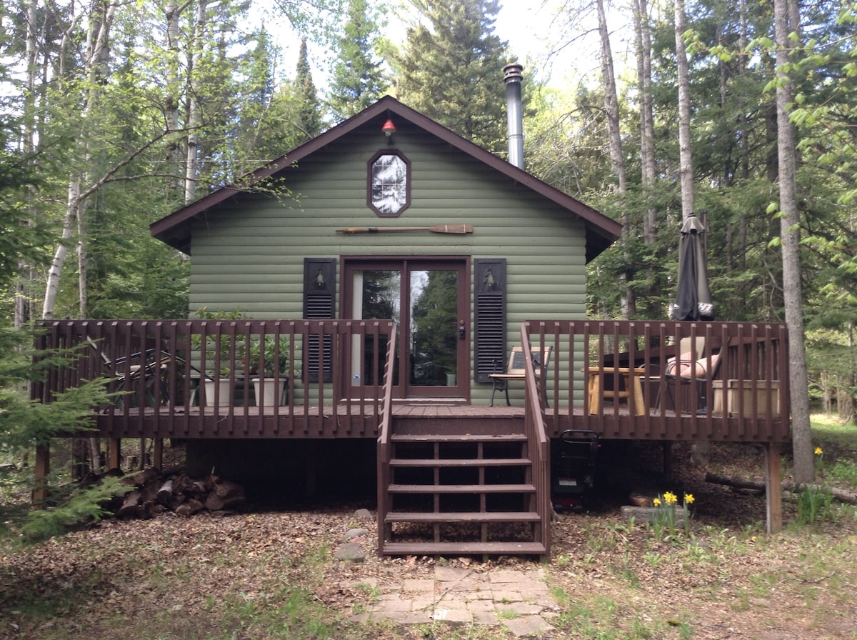 The cabin features a green exterior with a large front deck that includes steps leading up to it. Seating arrangements are set on the deck, surrounded by a wooded landscape. A wooden beam and a wood stove chimney are visible, with a small grill seen on the ground below.