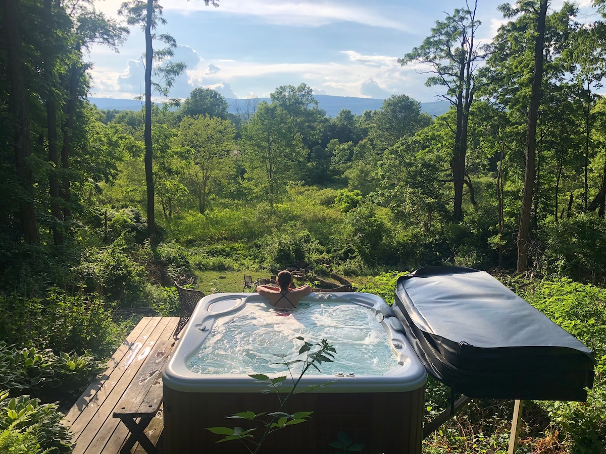 A hot tub is situated on a wooden deck, offering views of lush greenery and rolling hills in the background. Tall trees surround the area, creating a private retreat. The clear sky above hints at a serene atmosphere, perfect for relaxation.