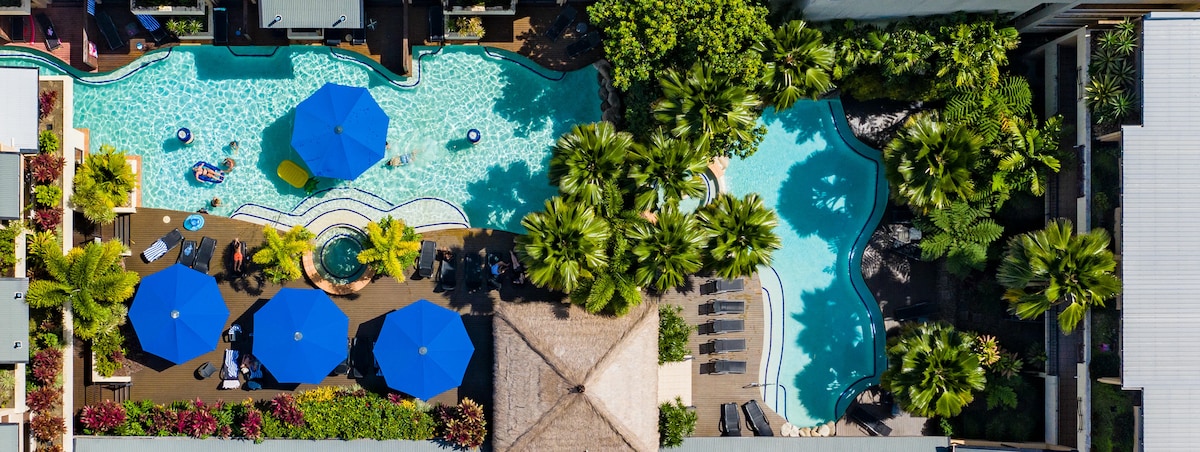 An aerial view showcases a sprawling pool area surrounded by lush greenery. Two saltwater pools are featured, adorned with blue umbrellas providing shade, and sun loungers are visible nearby. Vibrant plant life complements the inviting outdoor space.