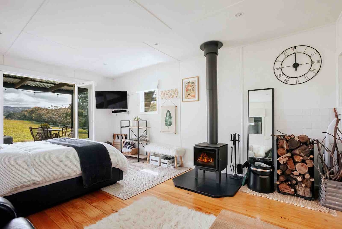 A cozy interior space showcases a queen bed layered with soft bedding and a plush throw. A sleek wood-burning stove stands prominently, accompanied by a stack of firewood. Large glass doors open to reveal scenic views of the rolling hills, while natural light fills the room.