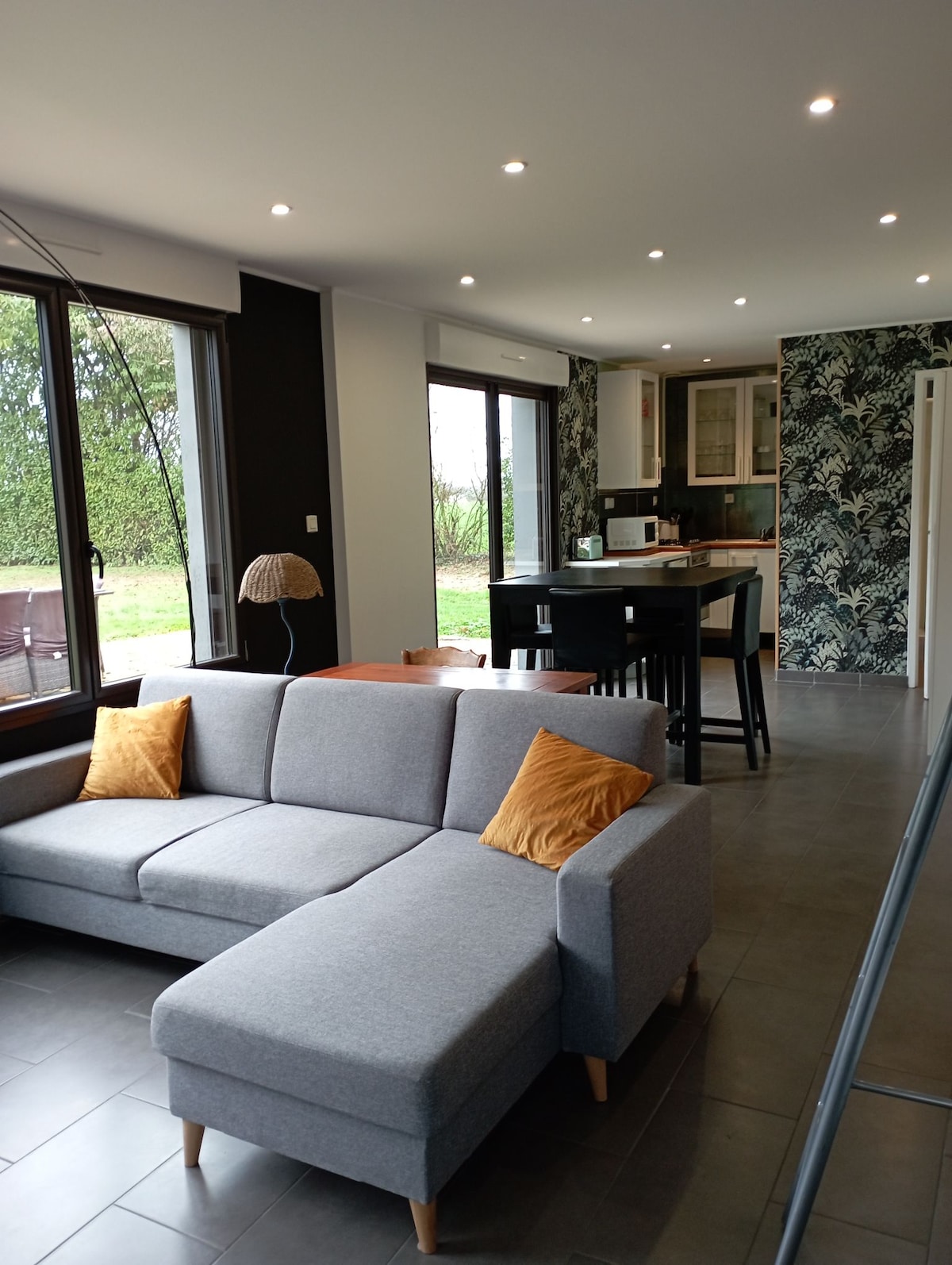 A modern living space is highlighted by large windows allowing natural light to fill the room. A gray sectional sofa with orange cushions faces a dining area and a sleek kitchen. The walls feature a contrasting dark color, while greenery is visible outside.