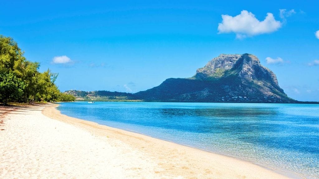 A sandy beach stretches along a calm coastline, with clear blue waters lapping gently at the shore. In the distance, a prominent mountain rises against a bright blue sky, creating a scenic backdrop. Lush greenery borders the beach, enhancing the natural surroundings.