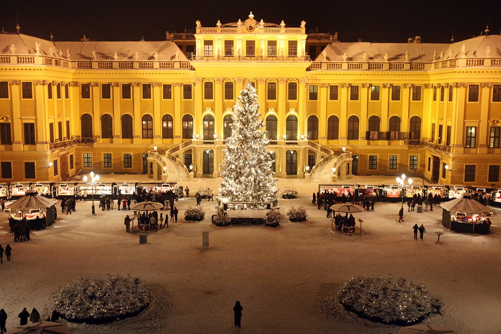 A beautifully illuminated courtyard is filled with snow, highlighting the grandeur of the surrounding architecture. A tall, decorated Christmas tree stands at the center, surrounded by market stalls and shoppers. Warm lights reflect off the building's facade, creating a festive atmosphere.