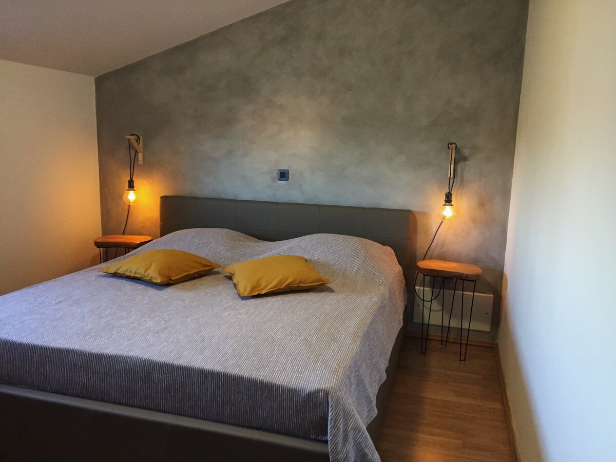 A spacious bedroom features a large bed flanked by two bedside tables with pendant lights. Soft yellow pillows add warmth, while the textured gray wall creates a calming backdrop. Natural light filters in, enhancing the serene atmosphere of the room.