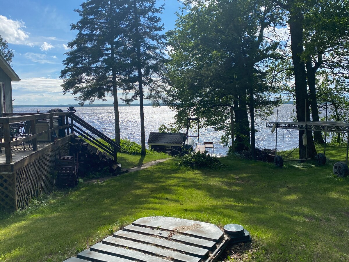 A view of the serene lake is captured through a cluster of trees. Sunlight reflects off the water, and a wooden structure is visible among the greenery. The scene presents a tranquil outdoor space perfect for relaxation and nature appreciation.