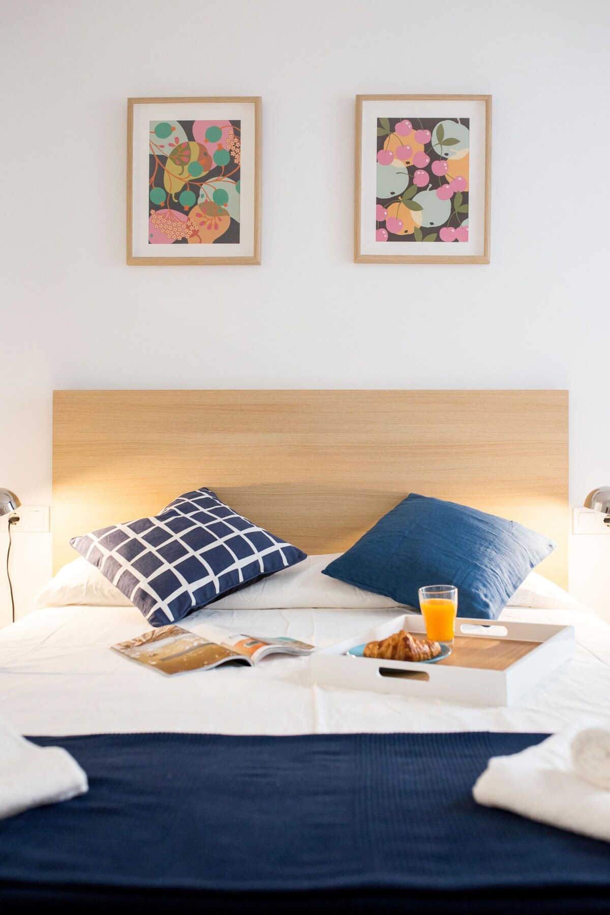 A double bedroom features a wooden headboard with decorative cushions placed at the back. Two framed artworks hang above the bed. A tray rests on the bed, holding a pastry and a glass of orange juice, alongside a magazine.