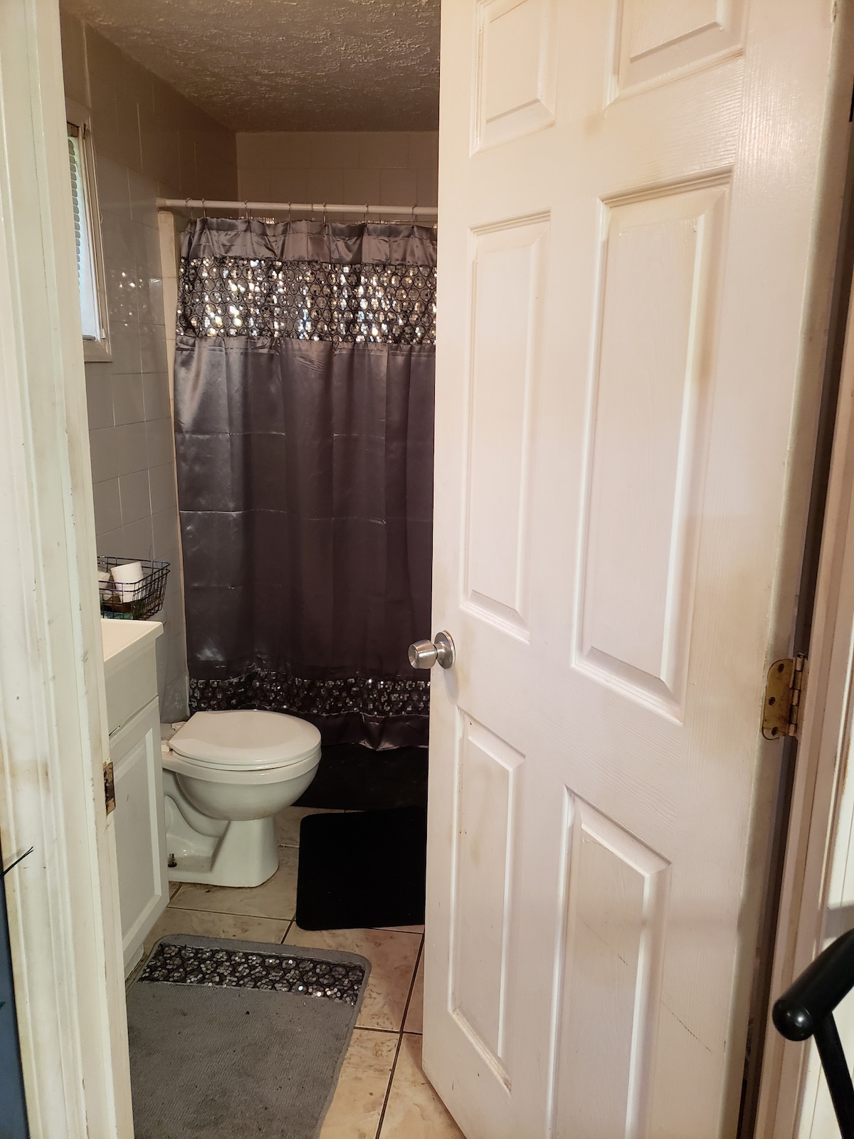 The bathroom is accessed through a white door, revealing tiled flooring and a shower area with a gray and silver curtain. A toilet is positioned next to a vanity with storage, and a gray bath mat lies on the floor.