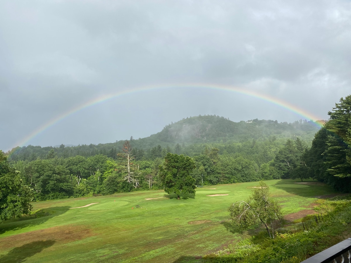 A lush green golf course is visible below, framed by rolling hills and trees. A vibrant rainbow arcs gracefully across the sky, connecting the landscape. The scene captures the tranquil beauty of nature in a serene, misty atmosphere.