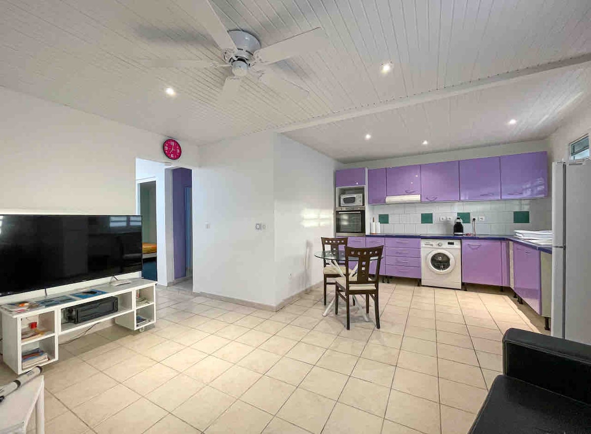 An open-plan kitchen and living area features light-colored tile flooring and white shiplap ceilings. Purple cabinetry contrasts with white appliances. A dining set with four chairs is positioned near a large flat-screen television, while sunlight enters through a nearby window.