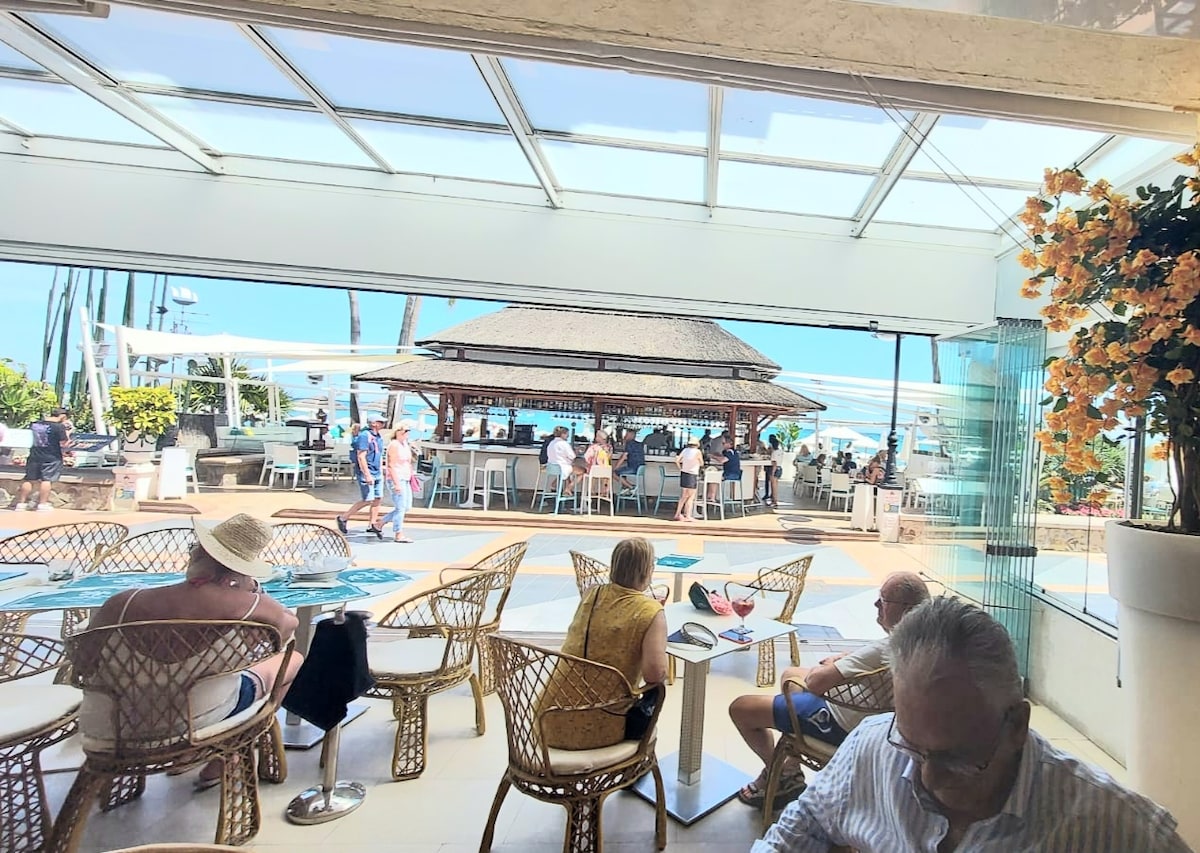 An open and airy space features woven chairs and a bar area with a thatched roof. Guests gather at tables while enjoying views of palm trees and distant beach. Natural light floods the area, enhancing the laid-back atmosphere of this social setting.