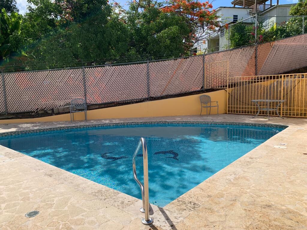 A bathing pool is surrounded by a textured patio, featuring a handrail for easy access. Lounge chairs and a table are positioned nearby, offering space for relaxation. Lush greenery and colorful flora are visible in the background, creating a serene setting.