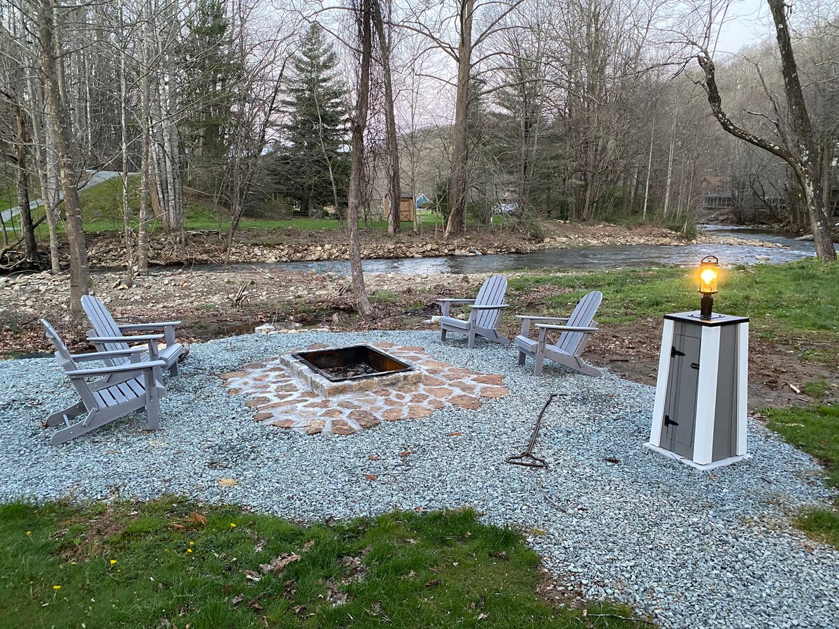 An outdoor gathering area features four Adirondack chairs arranged around a stone fire pit, set on gravel. A classic lamp provides soft lighting nearby, with the peaceful Watauga River flowing in the background and surrounded by trees.