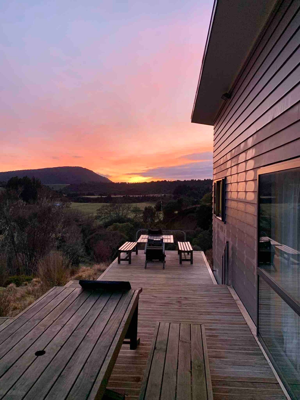 A wooden outdoor deck featuring a dining table set for six faces the sunset, with expansive views of rolling hills and native bush. The house's modern facade showcases large windows, reflecting the vibrant colors of the evening sky.