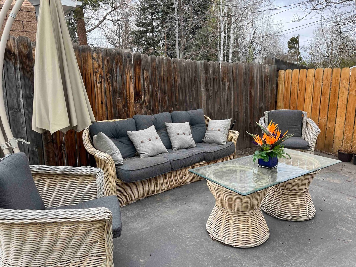 A private outdoor space features a comfortable seating arrangement with two wicker chairs and a cozy sectional sofa, all adorned with pillows. A glass-top table sits at the center, complemented by a vibrant floral centerpiece. A wooden fence encloses the area, enhancing privacy.