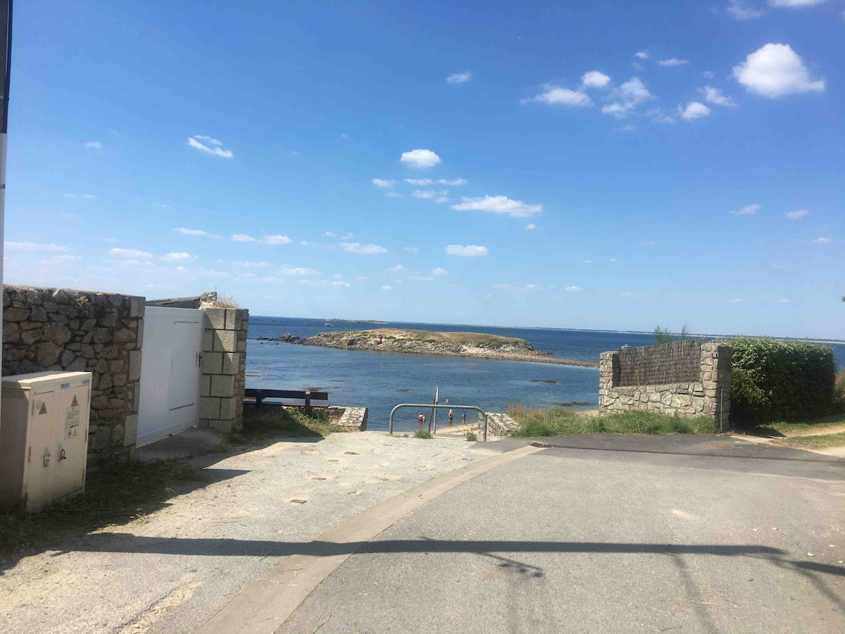 A scenic view of the ocean is presented, with calm waters reflecting the blue sky dotted with clouds. A rocky island sits in the distance, framed by a gravel pathway leading to the shore, highlighting the serene coastal environment.