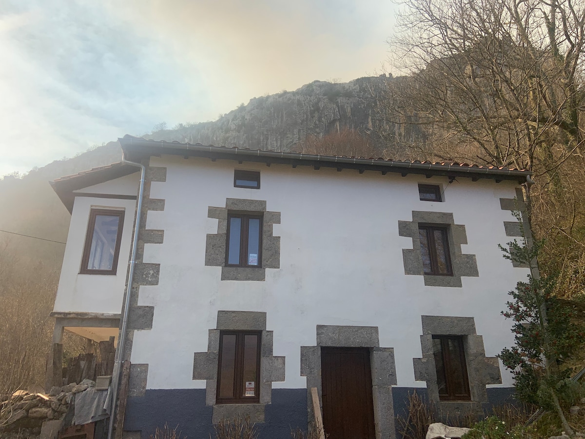 A charming house is displayed with a white facade and dark stone accents. The structure features several windows, allowing natural light to enter. A mountainous backdrop is visible, enhancing the serene setting of the location.