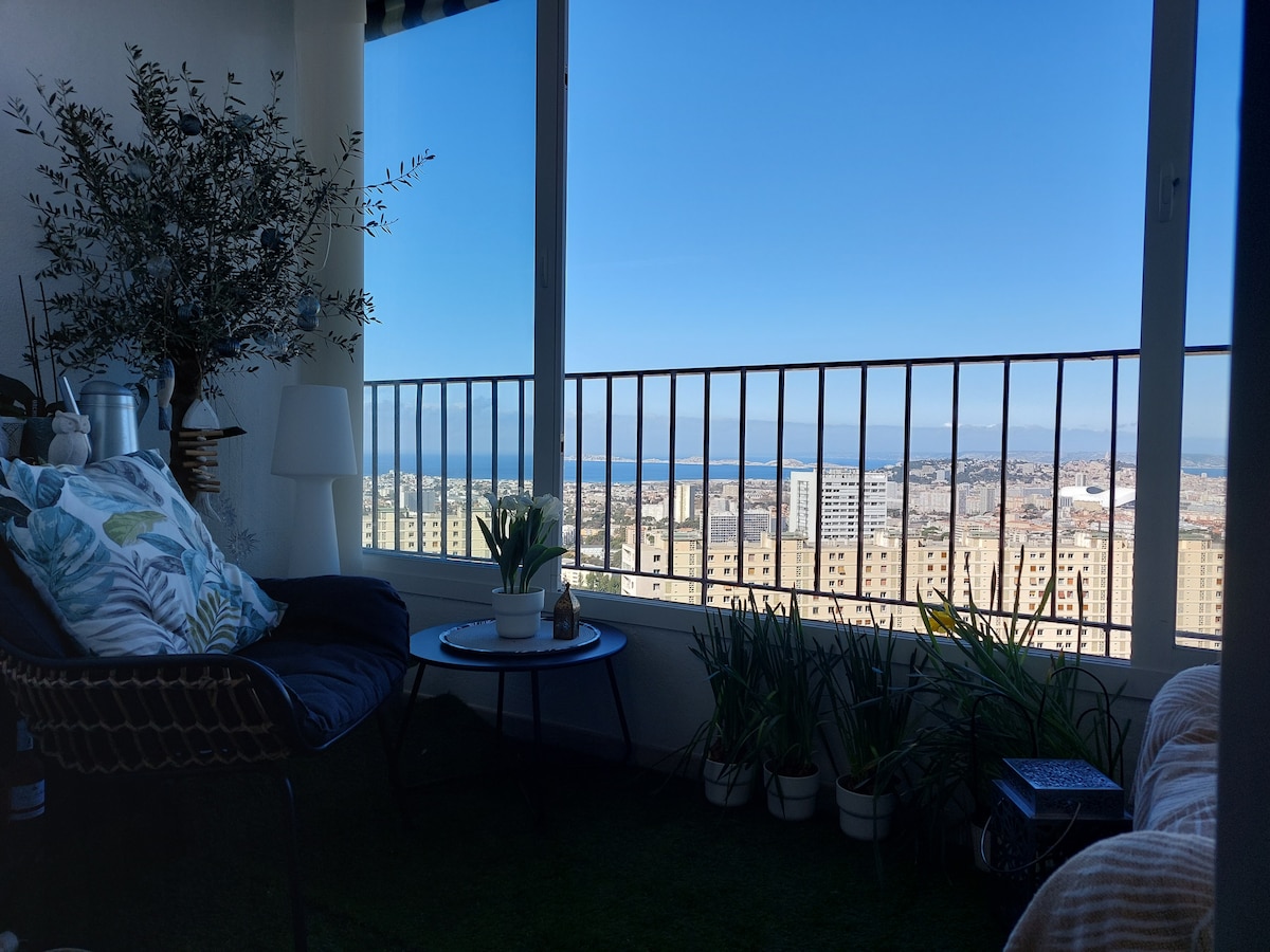 A cozy seating area is positioned next to large windows, offering expansive views of the sea and cityscape. Potted plants and decorative items enhance the space, while a small round table is set nearby. Natural light fills the area, creating a bright and airy ambiance.
