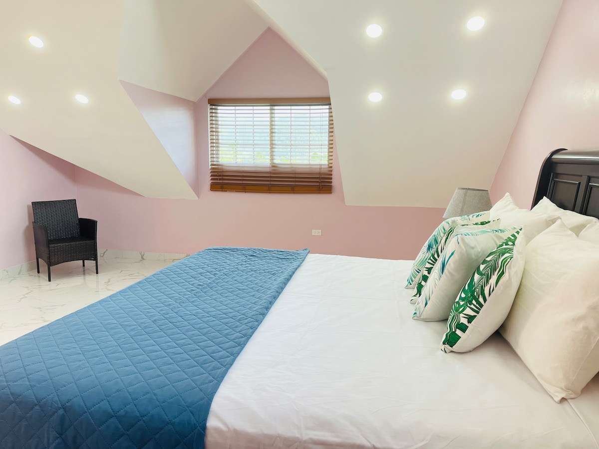 A spacious sleeping area features a large bed with decorative throw pillows against a backdrop of soft pink walls. A cozy chair is positioned in the corner, and natural light filters through the window adorned with wooden blinds.