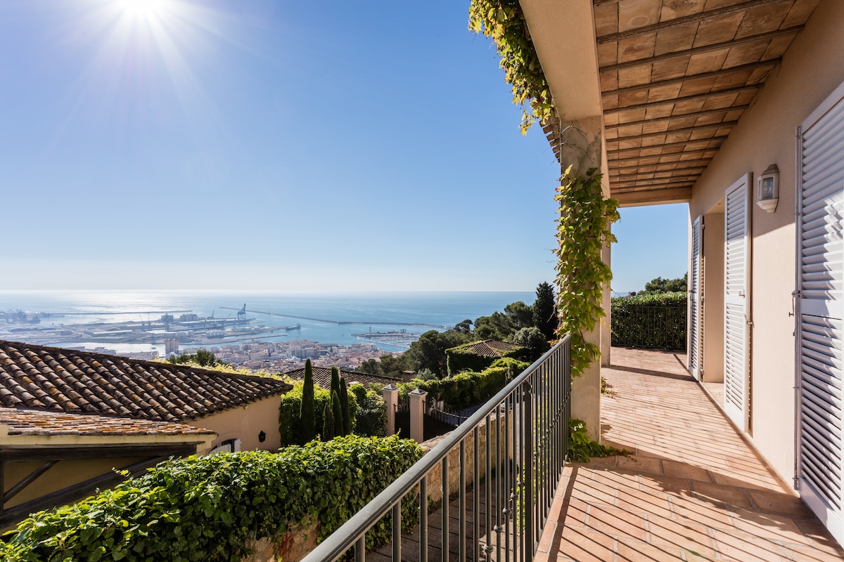A terrace overlooks the city and port of Sète, featuring a railing adorned with climbing plants. The expansive view showcases the sea and nearby landscape under a bright sky, with sunlight casting a warm glow on the surrounding area.