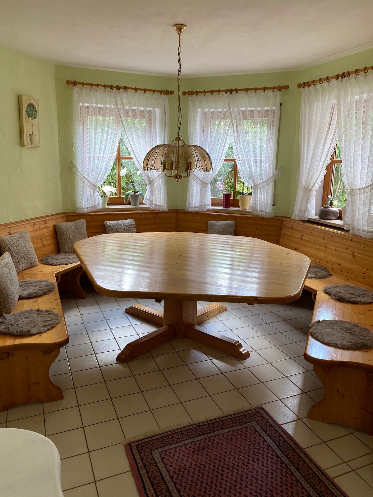 A spacious dining area features a large, oval wooden table surrounded by cushioned bench seating. Natural light filters through lace curtains on two windows, illuminating the warm tones of the wooden accents and creating a cozy environment.
