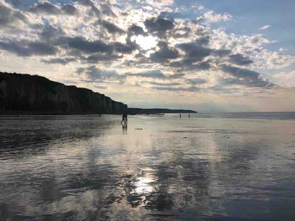 A calm beach scene is displayed, with soft waves lapping at the shore. Silhouettes of people can be seen walking along the wet sand, reflecting the sky's hues. The cliffs rise in the background, and a mix of dark clouds and sunlight creates an atmospheric sky.