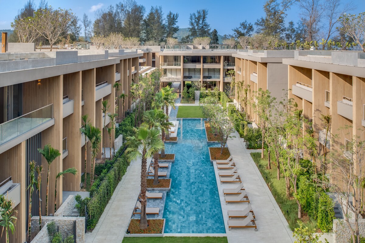 A tranquil resort courtyard features a long, narrow swimming pool flanked by lounge chairs and lush tropical landscaping. The area is surrounded by modern buildings with balconies and large windows, creating a serene atmosphere enhanced by palm trees and manicured green lawns.