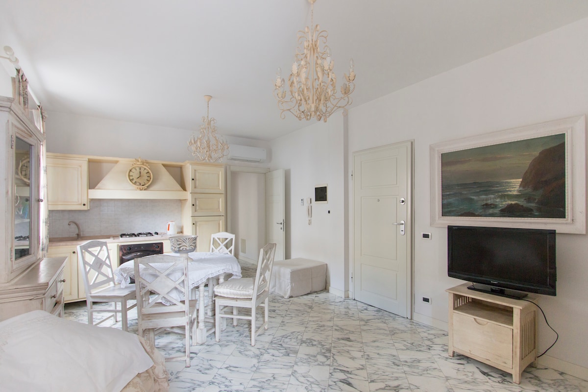 A bright living space features elegant chandeliers and white marble flooring. A light wood dining table with chairs is positioned near a cozy kitchen area, which includes cabinetry and essential appliances. A TV is mounted on the wall, and a landscape painting adds a decorative touch.
