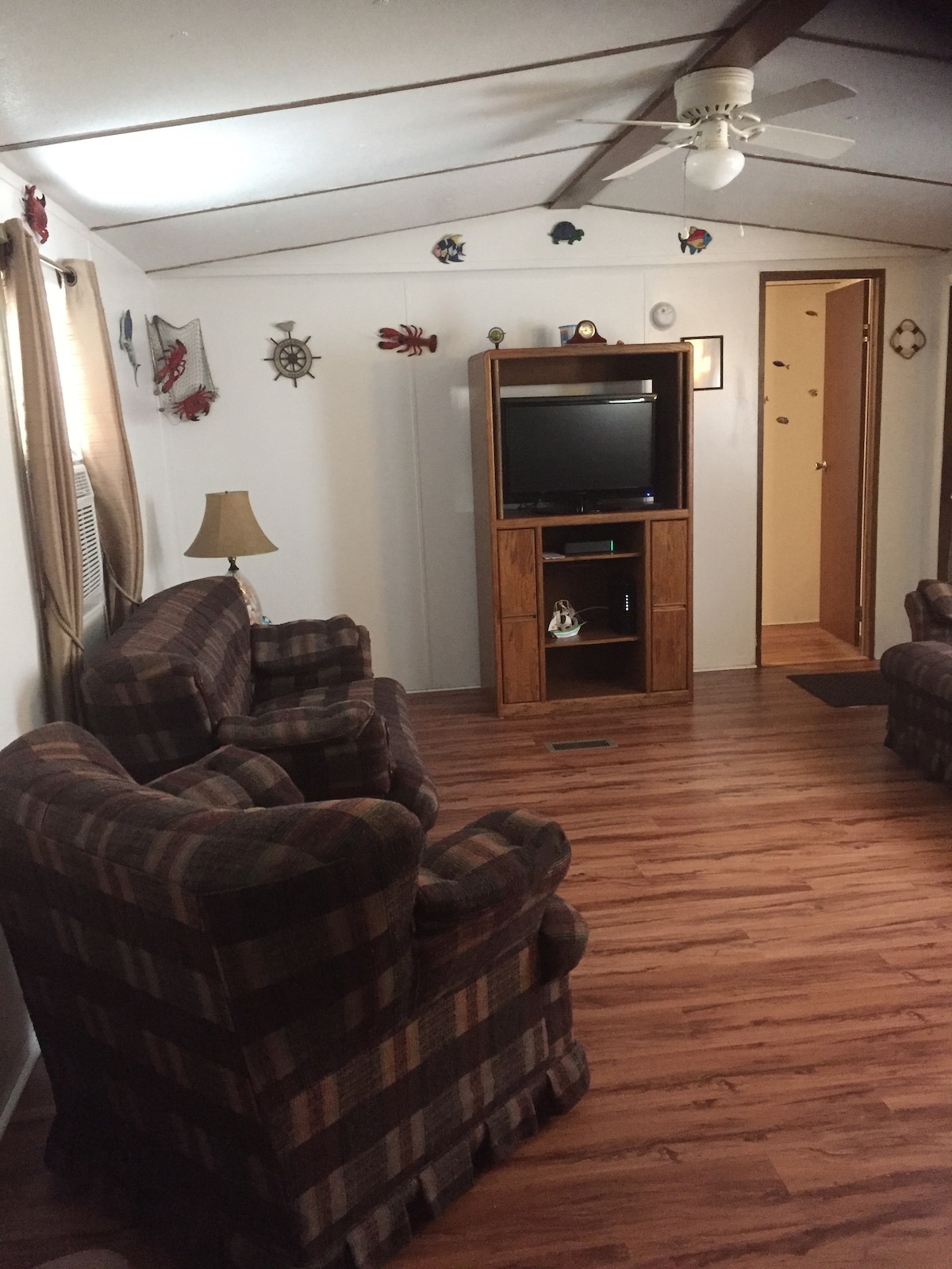 A living area features two plaid armchairs and a wooden entertainment unit with a television. Wall decorations add character, while natural light enters through the nearby windows covered by beige curtains. The flooring is finished in a warm wood tone, creating a cozy space.