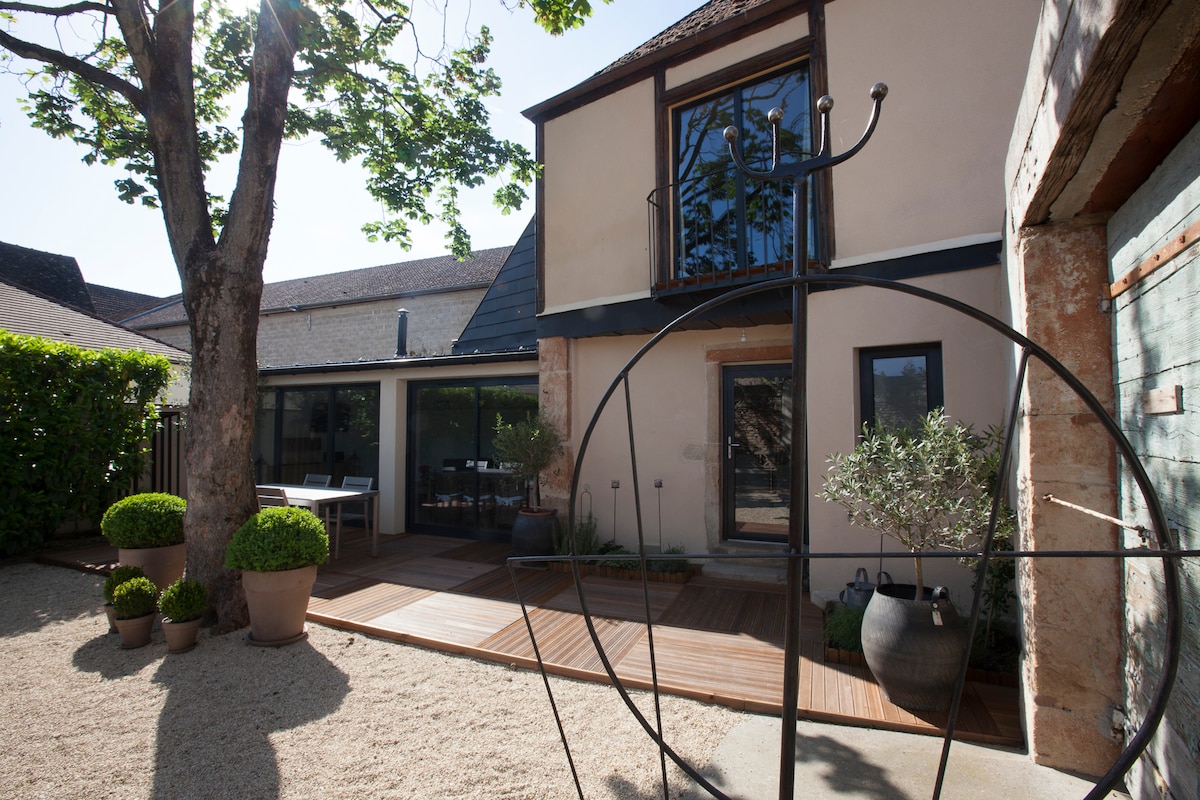 The exterior of the house is visible, showcasing a mix of natural stone and modern architecture. A wooden deck leads to the entrance, surrounded by landscaped greenery. A sculptural element is featured in the foreground, contributing to the tranquil outdoor setting.