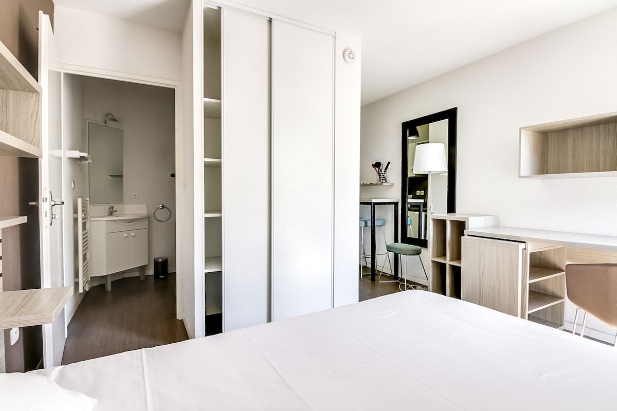 The room features a spacious bed with crisp white linens. A desk and chair are positioned near a mirror, while a kitchenette area is visible in the background. A closet with sliding doors is present, along with an attached bathroom doorway.