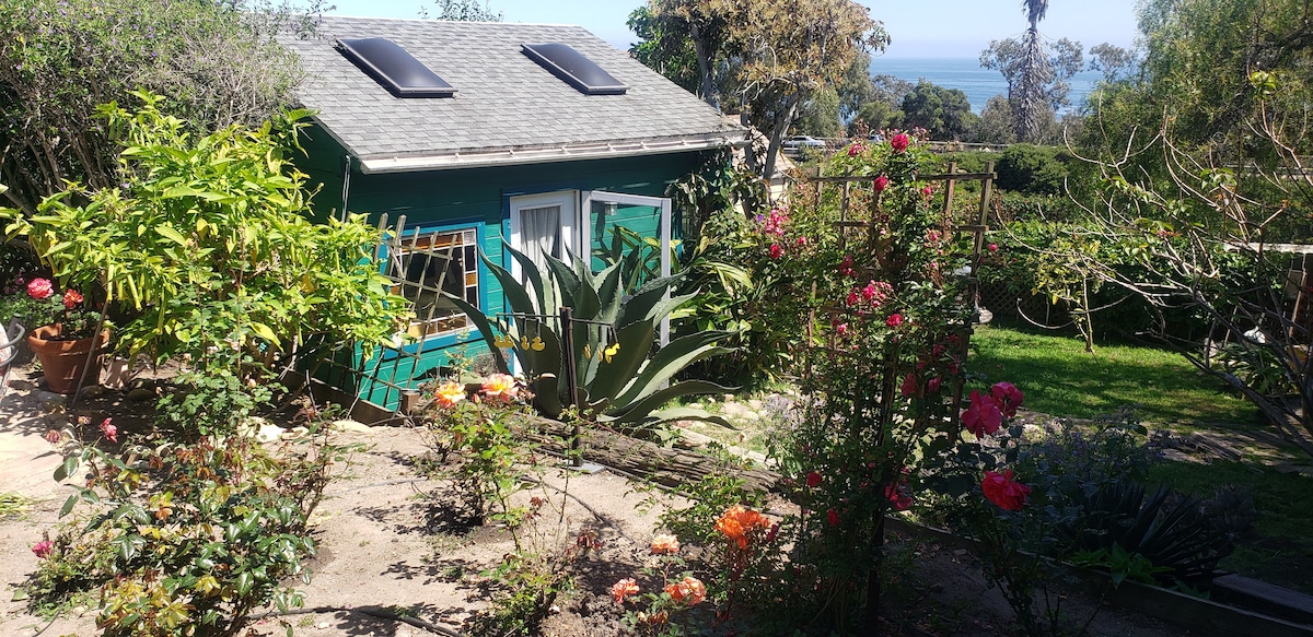 A charming garden studio is set amidst vibrant flowers and lush greenery. The exterior features a teal color with solar panels on the roof. Seaside views are visible in the background, framing the serene environment of the backyard.