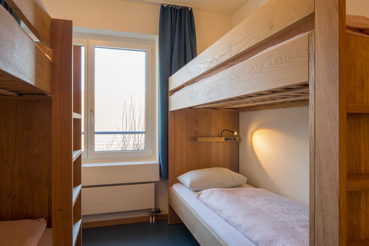 A well-lit bunk room features wooden bunk beds with ample space below. Soft bedding is laid out on each level. A window allows natural light to flow in, framed by dark curtains, creating a bright yet cozy atmosphere.