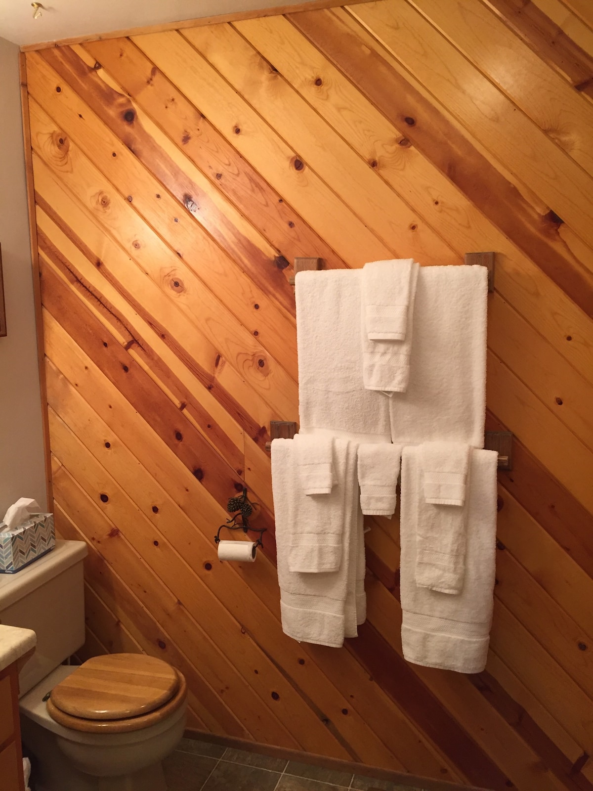 A bathroom features warm wooden paneling that creates a cozy atmosphere. White towels are neatly hung on a rack, with a matching tissue box placed nearby. A toilet with a wooden seat is visible, complementing the natural wood tones.
