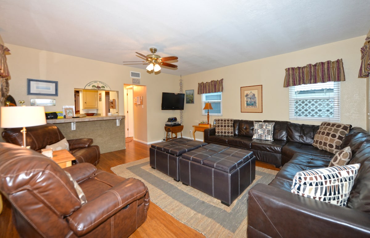 The spacious living room features two comfortable leather sofas and a large tufted ottoman at its center. A ceiling fan provides airflow, while light enters through windows adorned with patterned curtains. A flat-screen TV is mounted on the wall, enhancing the overall setting.