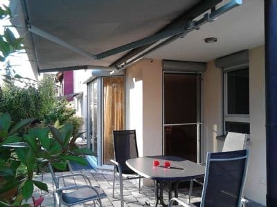 A covered terrace area is presented, featuring a round table with red accents and metal chairs. Lush greenery surrounds the space, creating a cozy outdoor setting. Large glass doors allow natural light to flow in while providing access to the indoors.