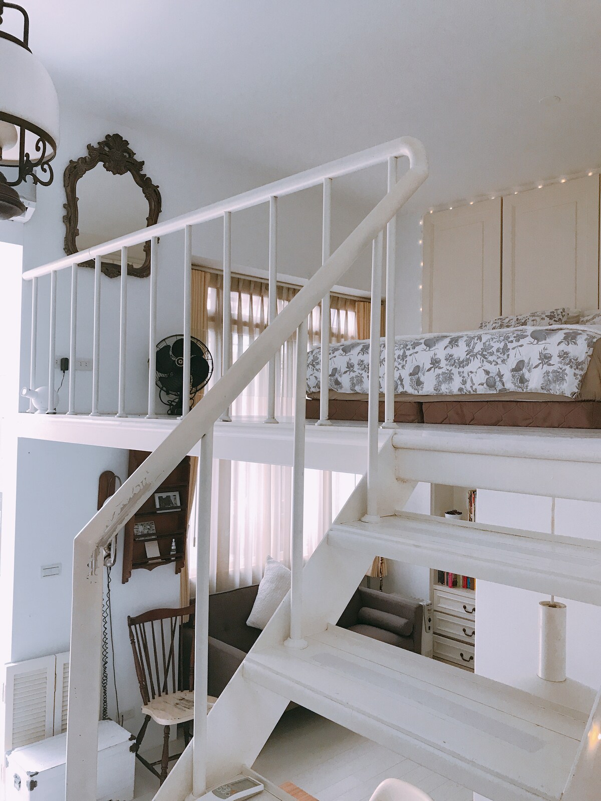 A white wooden staircase leads to an elevated sleeping area featuring a bed adorned with floral bedding. Natural light filters through adjacent windows, highlighting the simplicity of the room's design. A decorative mirror and a wooden chair add to the home's charm.