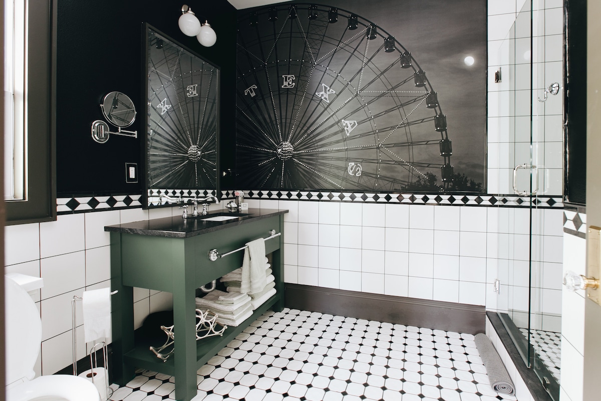 A modern bathroom features a striking black and white color scheme, highlighted by a large Ferris wheel mural. A dark green vanity is paired with white towels neatly arranged. The floor is adorned with black and white patterned tiles, complemented by a spacious glass shower.