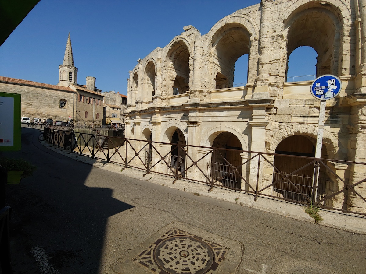 The image captures the iconic Arènes d'Arles, showcasing its ancient stone arches and intricately detailed facade. A tall spire of a nearby building rises in the background, along with visible street signs and a paved pathway lined with a wooden fence.