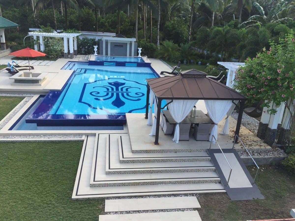 A large swimming pool with an intricate design occupies the center of the outdoor space. Surrounding the pool are multiple lounging areas, including a gazebo with white drapery. Lush greenery and palm trees frame the scene, enhancing the serene atmosphere.
