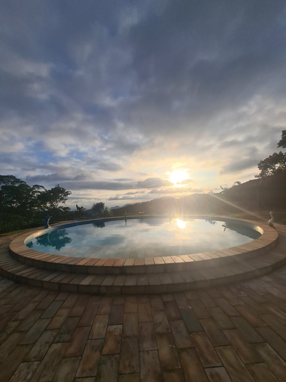 A round pool is set within a stone patio, surrounded by greenery. The water reflects the golden light of the setting sun, while clouds create a soft gradient in the sky. The tranquil scene is enhanced by distant silhouettes of trees.