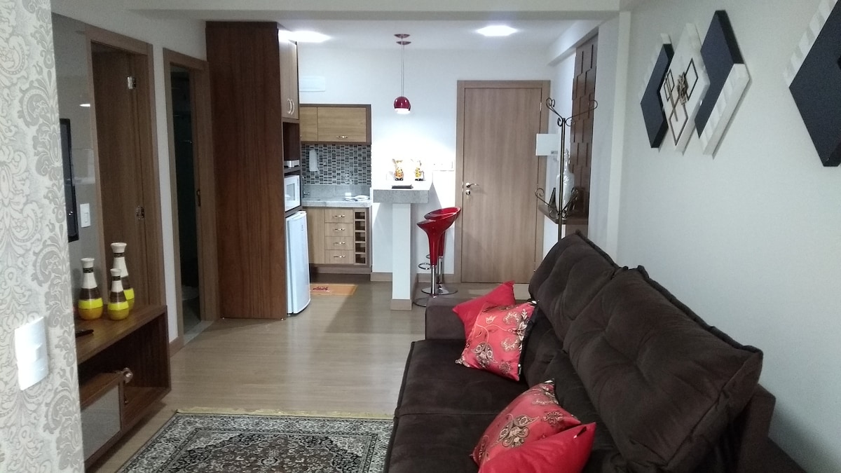 A living area features a dark brown sofa adorned with decorative pillows. The compact kitchen, visible in the background, includes wooden cabinets and a bar area with red stools. A woven rug and wall art complement the light-colored walls and wooden flooring.