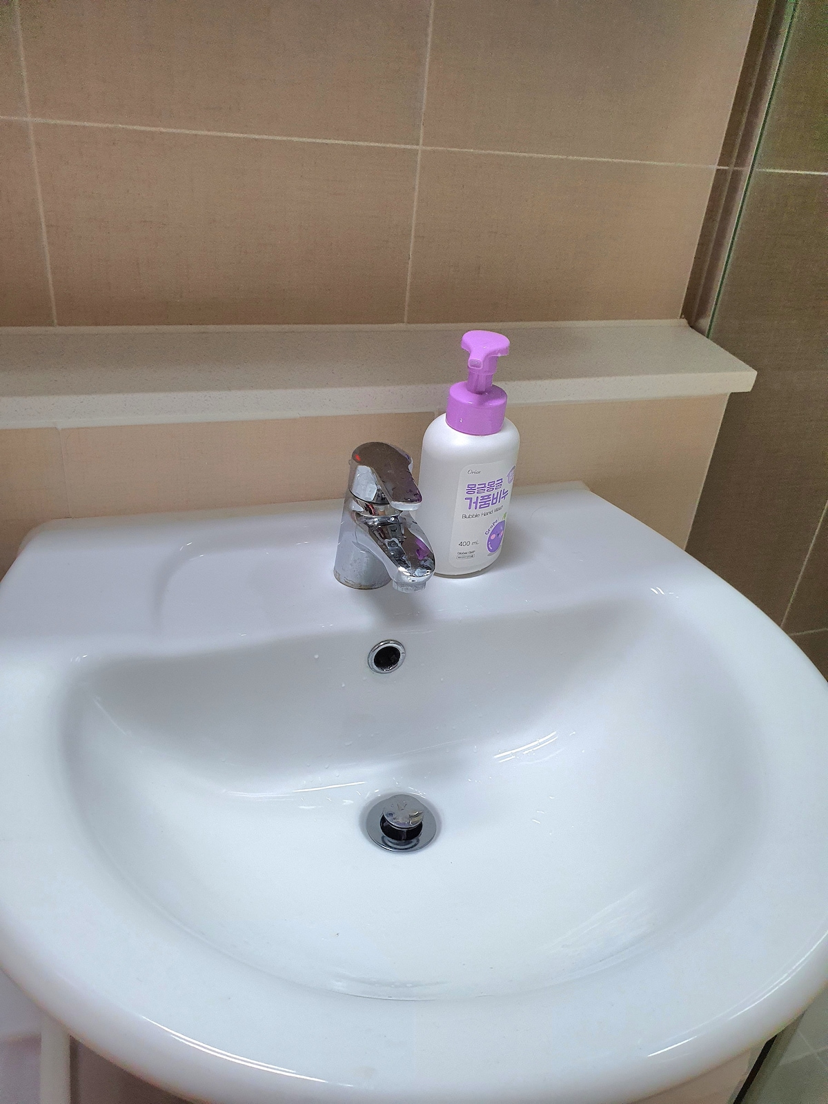A modern sink is seen with a smooth, white ceramic basin and a polished chrome faucet. A bottle of hand soap with a pink pump lid is placed beside the sink, resting on a solid counter surface. Neutral tiled walls complement the overall clean design.