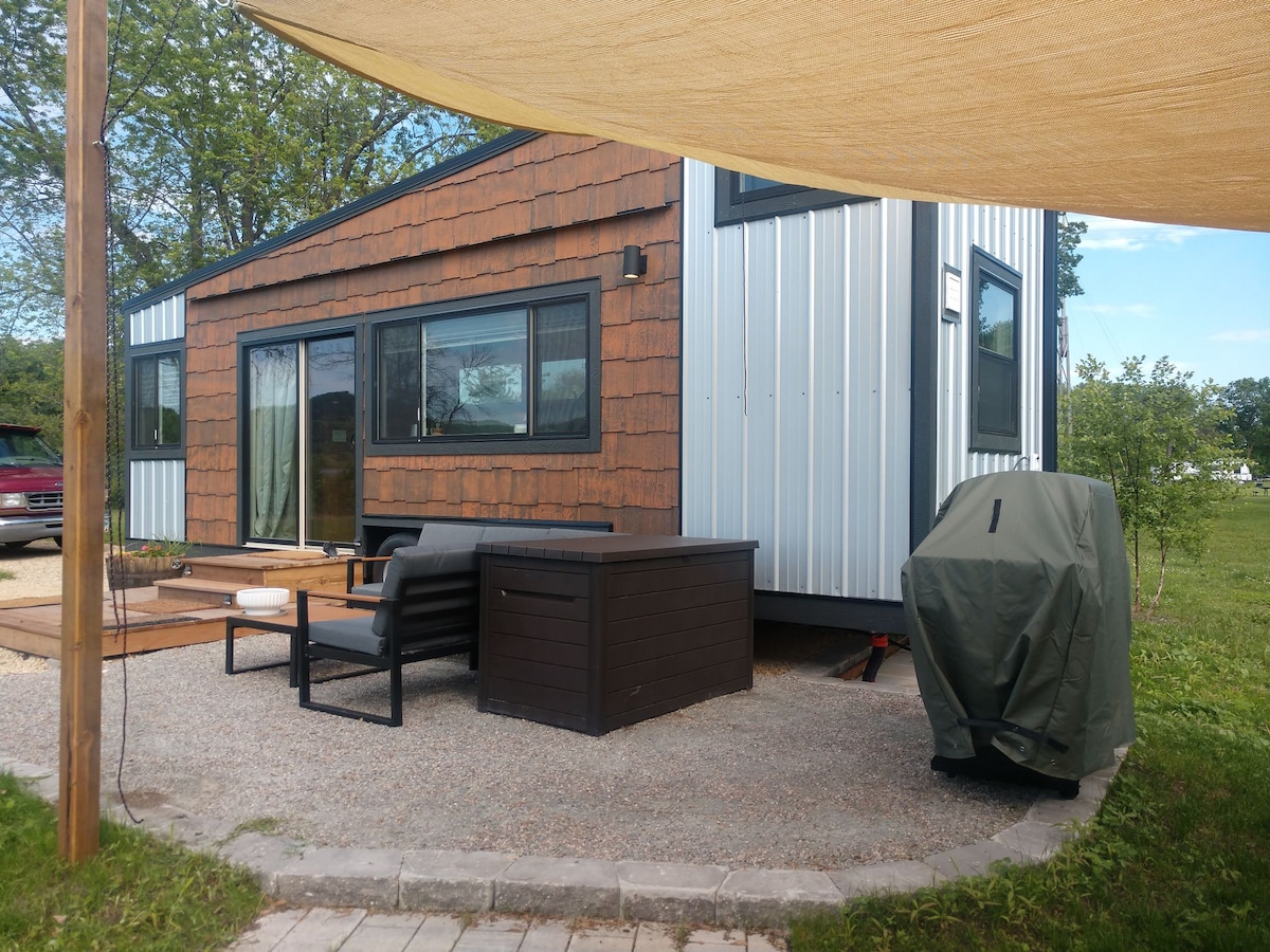 The exterior of a contemporary tiny house is shown, featuring a mix of wood and metal siding. A small patio area includes a black seating arrangement and a storage box. A covered grill area is visible nearby, surrounded by grassy landscaping.