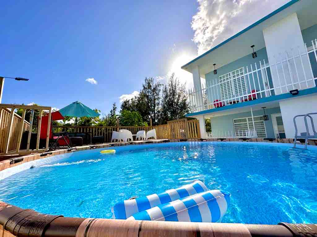 A circular pool is surrounded by a wooden deck, with two lounge floats resting on the water's surface. Sunlight glimmers off the clear, inviting blue water. A shaded area with an umbrella and seating is visible in the background, offering a relaxing outdoor space.