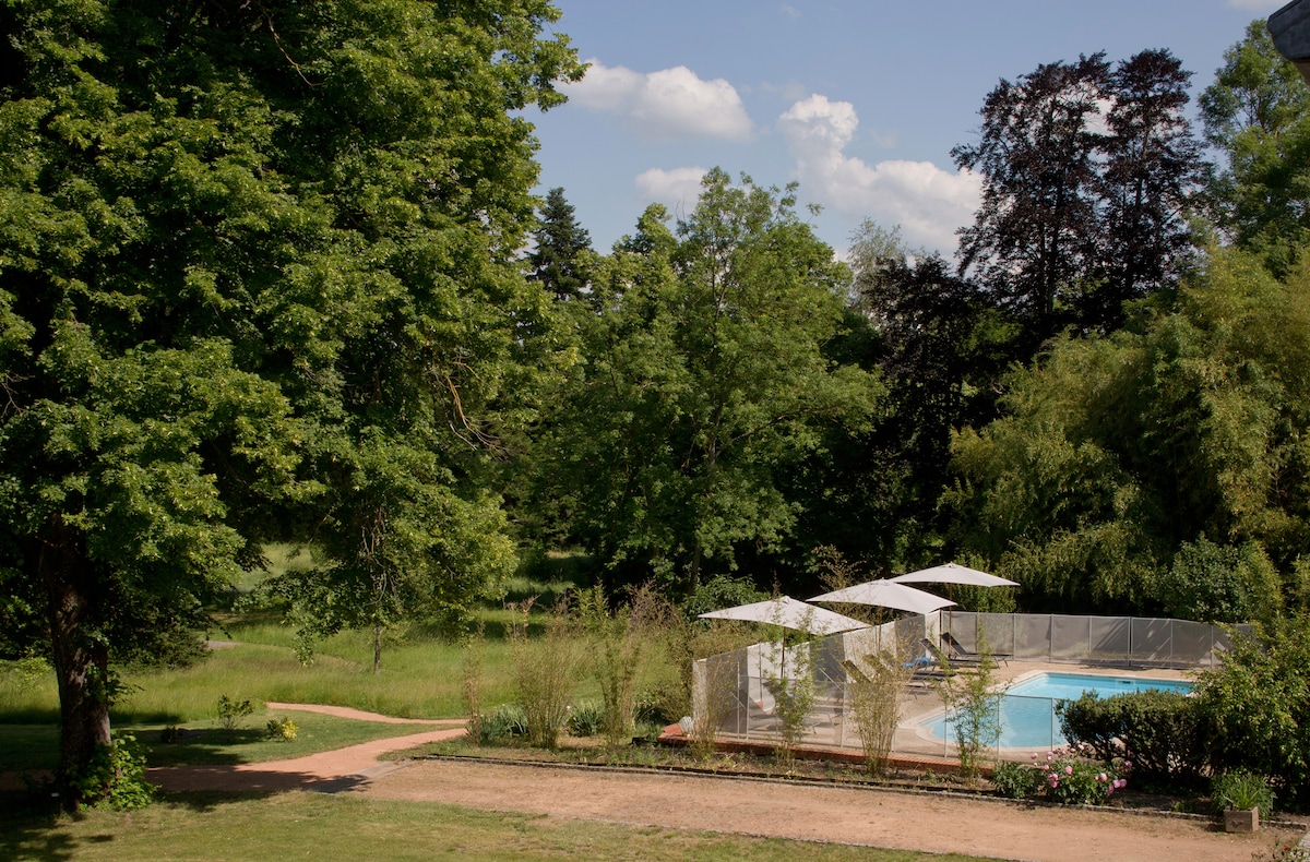 A shared outdoor pool is surrounded by greenery, featuring protective fencing for safety. Sun loungers with umbrellas are visible beside the pool. Lush trees and plants fill the surrounding area, creating a serene environment for relaxation.